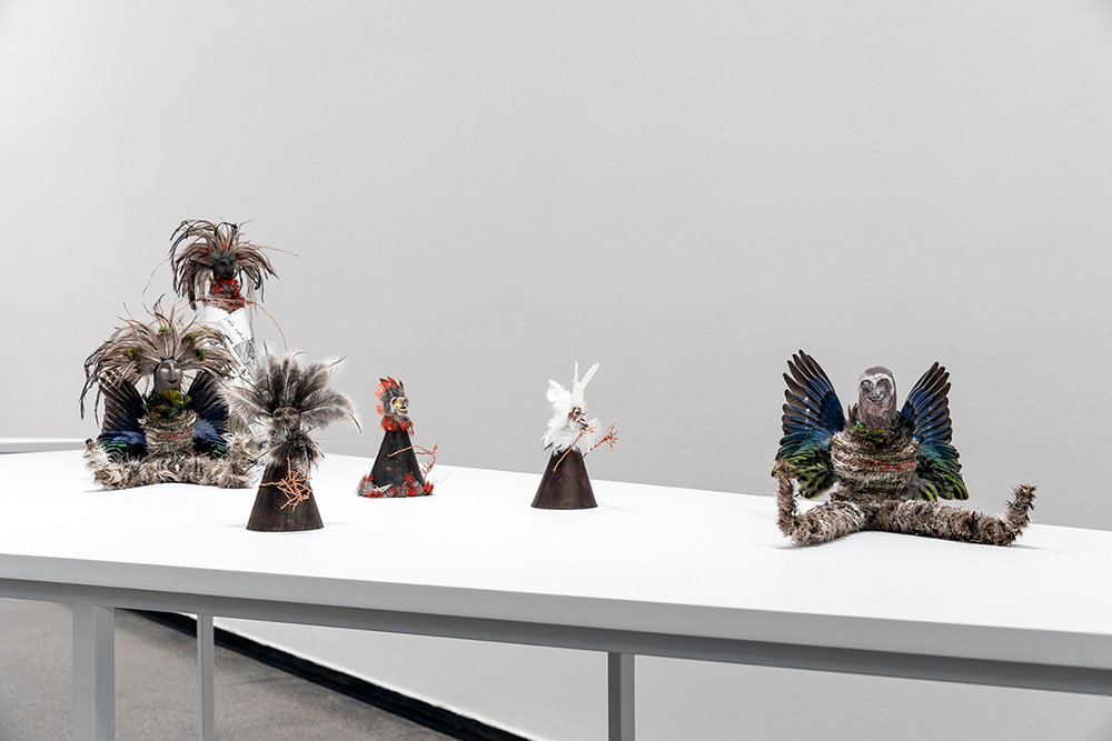 Lisa Waur, Ancestors (Detail), 2018, 2 cloaked figures: feathers, ceramic, glaze, digital print on cotton, coper wire, birds feet and metal stands, 41 x 28 x 50 cm each, 2 winged figures: feathers, ceramic, glaze, oaten hay, fibre, bird’s wings, wool, coton, 30 x 30 x 20cm each. Family (Detail), 2018, 5 figures: feathers, ceramic, glaze, forged recycled water heater, copper, patina, copper wire, fibre, 20 x 13 x 13 cm each, Installation view, Australian Centre for Contemporary Art (ACCA), 2018, Photo Andrew Curtis, Courtesy the artist and This is No Fantasy + Dianne Tanzer Gallery Melbourne