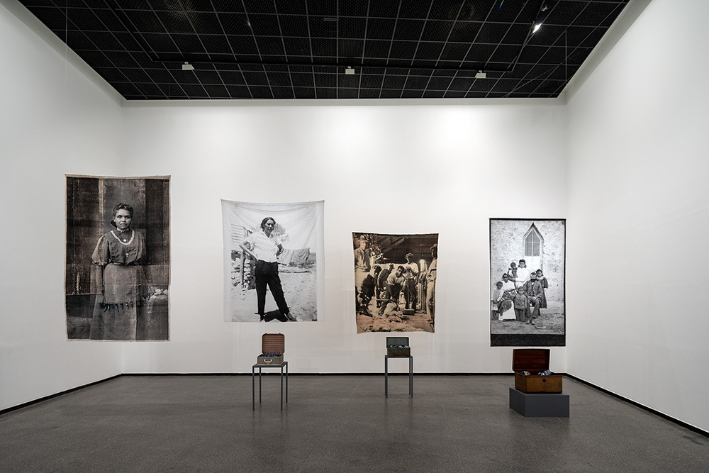Yhonnie Scarce, from the series Remember Royalty, 2018: Granny Melba, 2018, Water based ink screen printed on vintage French linen, black lustre blown glass, 300 x 191 cm; Fanny – Andamooka opal fields, South Australia, 2018, Water based ink screen printed on vintage cotton sheet, vintage suitcase, black lustre blown glass, vintage gloves, embroidered handkerchiefs sheet: 260 x 230 cm, suitcase with objects: 62 x 47 x 50 cm; Papa Willy, 2018, Water based ink screen printed on woollen blanket, black lustre blown glass, tool box, metal hand tools blanket: 235 x 202 cm, toolbox with objects: 41 x 50 x 27 cm, Family portrait – Koonibba Mission, South Australia, 2018, Water based ink screen printed on vintage cotton sheet, vintage metal trunk, black lustre blown glass sheet: 258 x 165 cm, trunk with objects: 33 x 72 x 54 cm; Installation view, Australian Centre for Contemporary Art, Photo: Andrew Curtis, Print production: Spacecraft, Collingwood Courtesy the artist and This is No Fantasy + Dianne Tanzer Gallery-Melbourne