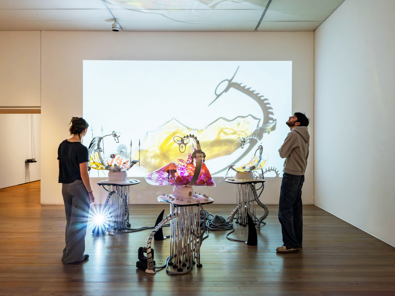 Katja Novitskova, Pattern of Activation (Mamaroo nursery, dawn chorus) Date of origin: 2017, Installation view Labouring Bodies at Museum Tinguely, Basel, 2026, 3 electronic baby swings, 3 aluminium folding stands, plastic cable covers, lasers, epoxy clay, digital print on polyurethane resin, robotic bugs, cable ties, plastic toy parts, foam stress pills, elastic puffers, plastic drill plugs, silicone stress eggs, fossils, tree mushrooms, magnets, programmable rotating light heads, custom speakers, and sound installation (sound piece by Kareem Lotfy), video projection file (dimensions variable), electric cords, Dimensions variable, site specific, © Katja Novitskova, Courtesy the artist and Kraupa-Tuskany Zeidler, Berlin/Munich, Photo Credit: 2026 Museum Tinguely, Basel; Pati Grabowicz