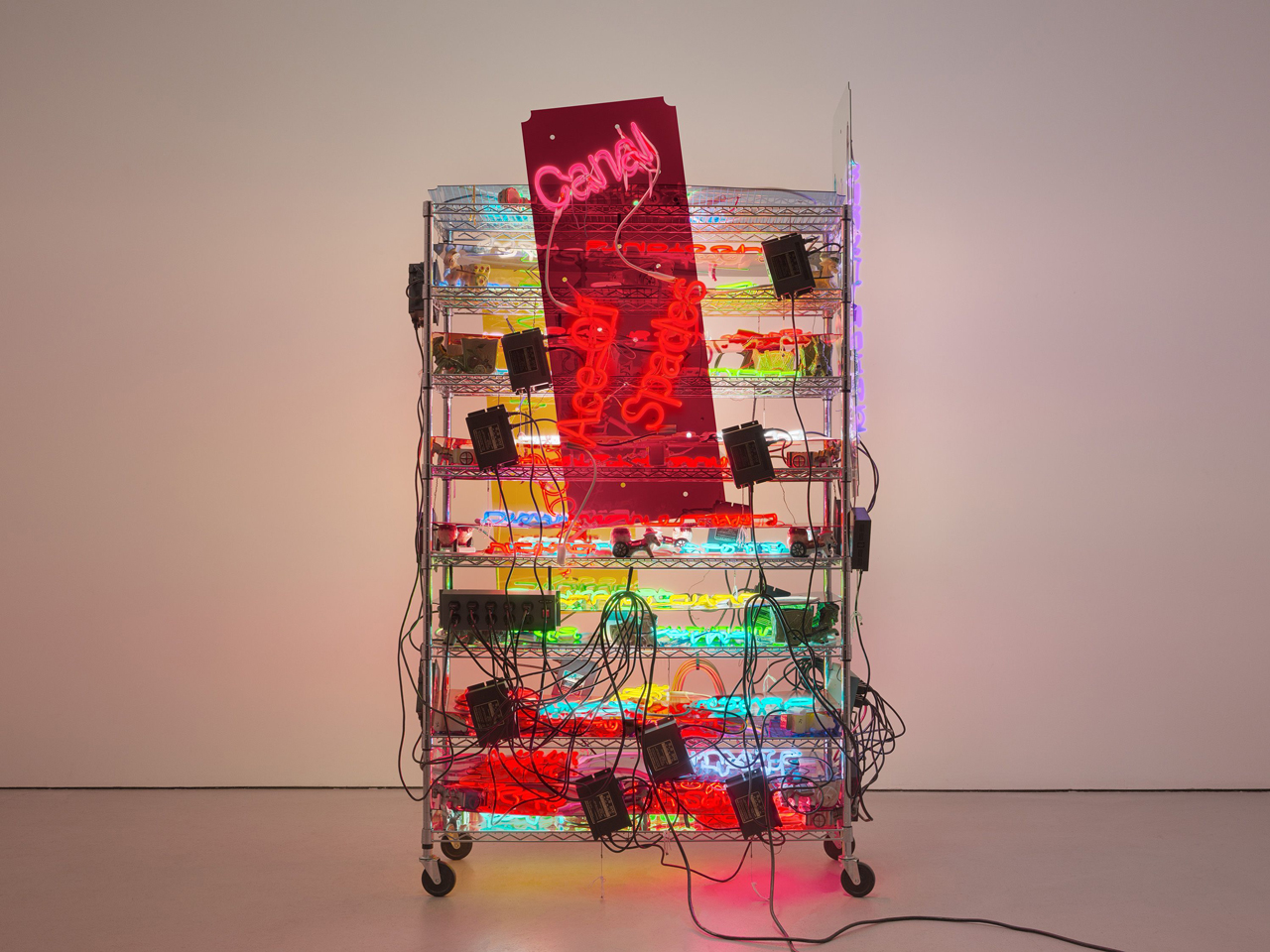 Jason Rhoades , Shelf (Ace of Spades), 2003, Eight-shelf rack on wheels, nineteen (19) Plexiglas panels with mounted neon-phrases, fifty-one (51) ceramic donkey carts, nineteen (19) 110V transformers, black power cord, and power strip. 92 1/2 x 54 x 27 1/2 inches (235 x 137.2 x 69.9 cm), © Jason Rhoades, courtesy the artist and David Zwirner Gallery