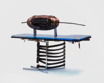 A blue acrylic tabletop resting on a metal chair with leather straps. A brown ovoid shape with a large stick behind it sits atop the table